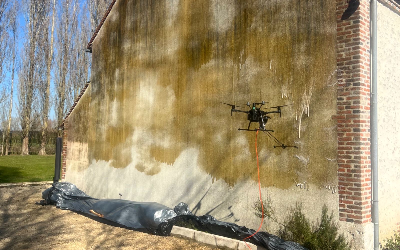 Drone BatiWash appliquant le traitement sur un pignon encrassé, abords protégés par des bâches